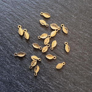 May include: Gold-coloured leaf charms with a textured surface and a small loop for attaching to jewellery. The charms are scattered on a dark, textured surface. These charms are ideal for DIY jewellery making.