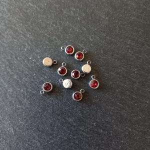 May include: A collection of small, circular silver-toned charms, each featuring a single, faceted deep red gemstone set in the centre. Each charm includes a small metal loop at the top for attachment, with some charms showing the plain silver back.