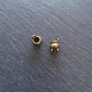 Peut inclure: Deux petits breloques dorées antiques. L'une est un chaudron à trois pieds et une poignée. L'autre est un petit cylindre à sommet ouvert avec une boucle. Les deux breloques sont posées sur un fond gris foncé texturé.