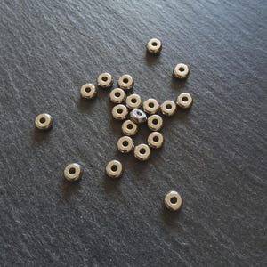 May include: A collection of small, shiny, silver-coloured metal beads with a circular shape and a hole in the centre. The beads are scattered on a dark grey, textured surface.