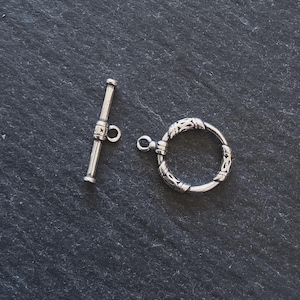 May include: A silver toggle clasp set, comprising a bar and a decorative ring. The bar has a small loop, and the ring is embellished with an intricate pattern. The clasp is designed for jewellery making.