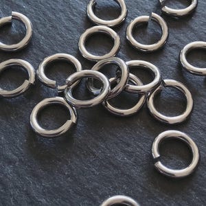 Op de afbeelding: Een verzameling glanzende, zilverkleurige sprongringen, verspreid over een donker, getextureerd oppervlak. De ringen zijn open, klaar voor gebruik in het maken van sieraden of andere ambachten. Het metaal heeft een gepolijste afwerking.