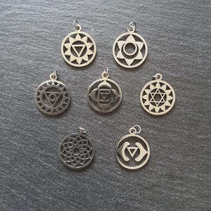 May include: Seven silver-colored chakra pendants with intricate geometric designs. Each pendant is circular, featuring symbols representing different energy centers. The pendants are displayed on a dark gray surface, showcasing their detailed craftsmanship.
