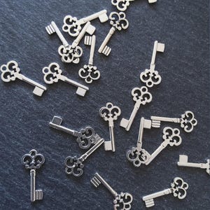 May include: A selection of small, silver decorative keys with elaborate, vintage designs. The keys are scattered on a dark, textured surface, highlighting their intricate details and classic style. Suitable for crafting or jewellery making.