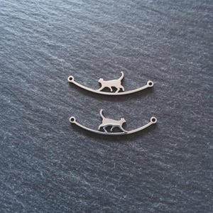 May include: Two silver cat charms. Each charm shows a cat silhouette walking along a curved line, with small holes at each end. The charms are displayed on a dark grey, textured surface.