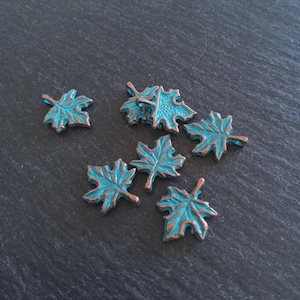 May include: Several small, leaf-shaped charms with a distressed teal and bronze finish. The charms are scattered on a dark gray surface. The leaves have detailed veining and a rustic appearance, suitable for jewelry making or craft projects.