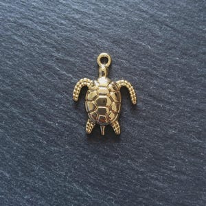 May include: A gold turtle charm, suitable for a necklace or bracelet, with a loop at the top. The turtle features a detailed shell and outstretched flippers. The charm is set against a dark, textured background.