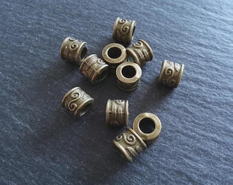 May include: A collection of antique bronze-coloured metal beads. Each cylindrical bead features a repeating scroll design. The beads are scattered on a dark grey surface, creating a textured contrast.
