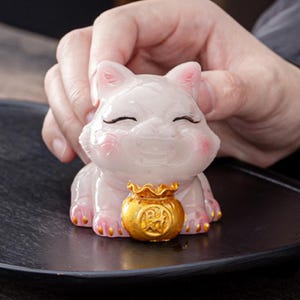 Könnte beinhalten: Eine kleine, blassrosa, durchscheinende Katzenfigur mit lächelndem Ausdruck, geschlossenen Augen und rosafarbenen Wangen. Die Katze hält einen glänzenden goldfarbenen Beutel mit dem chinesischen Schriftzeichen für Reichtum und hat goldene Akzente an den Pfoten.