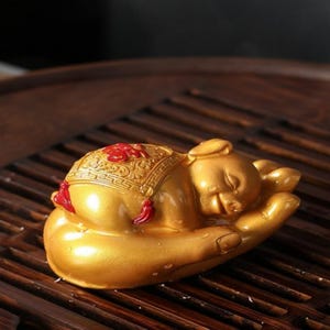 May include: A gold-colored figurine of a sleeping piglet resting in a cupped hand. The piglet wears a decorative saddle featuring intricate patterns and red accents, including a prominent red Chinese character on its back and small red tassels.