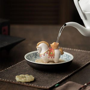 May include: A white ceramic horse figurine with gold accents and a saddle, placed in a decorative bowl with a blue and white pattern. Water is being poured from a white teapot into the figurine. The horse is on a brown surface.