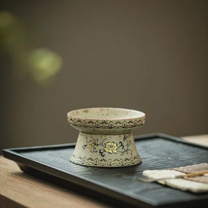 May include: A cream-coloured ceramic pedestal bowl with a wide, shallow top, decorated with yellow floral patterns and dark blue accents. The bowl sits on a black tray, suitable for a tea ceremony or decorative display. The bowl is likely for serving.