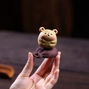 Peut inclure: Une petite figurine en céramique beige et ronde représentant une créature ressemblant à un ours avec des joues roses et une petite bouche, posée sur une base circulaire texturée brun foncé. La base présente des motifs circulaires en relief.