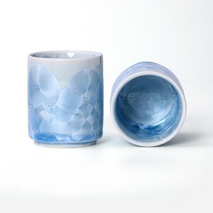 Peut inclure: Deux tasses en céramique avec un glaçage cristallin bleu clair et blanc. Une tasse est droite, l'autre est sur le côté, révélant l'intérieur. Les tasses ont une forme cylindrique et une base légèrement évasée.
