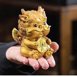 Könnte beinhalten: Goldene Figur einer Fabelwesen mit Flügeln, Hörnern und einem Schnurrbart, die eine Münze hält. Die Figur ist mit aufwendigen Designs versehen und sitzt in einer sitzenden Position. Die Gesamtästhetik ist verziert und dekorativ.