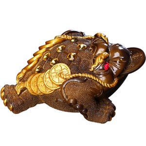 Op de afbeelding: Een bronzen, driepotige padfiguur met gouden accenten. De pad houdt gouden munten in zijn mond en is versierd met gouden juwelen. Een rode edelsteen is in het voorhoofd gezet. De figuur is een symbool van rijkdom en voorspoed.
