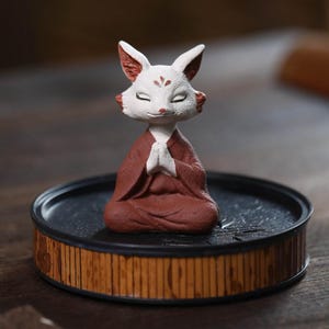 以下が含まれることがあります： 瞑想のポーズをとるセラミック製のキツネの置物。マルーン色のローブを着て、竹風の縁が付いた黒いトレイに座っています。キツネは白い毛皮で、耳と顔に赤いアクセントがあり、目は閉じられています。