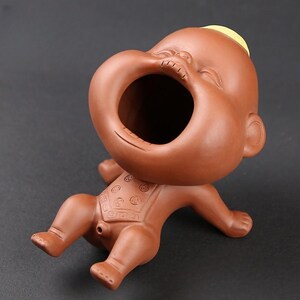Può includere: Statuina in ceramica marrone raffigurante un bambino con una grande bocca aperta. Il bambino è sdraiato sulla schiena con le braccia e le gambe distese. Ha una ciocca di capelli gialli sulla testa e un bavaglino fantasia.