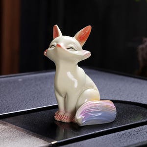 May include: A cream-coloured ceramic fox figurine in a seated pose with closed eyes. The fox has pink paws and orange-tipped ears. Its tail is a blend of pastel colours. The figurine is on a black surface.