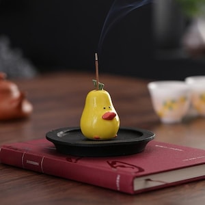 May include: A yellow pear-shaped ceramic incense holder with a small orange beak and green leaf detail, resting on a circular black tray. A thin brown incense stick is inserted into the top of the pear, emitting a faint wisp of grey smoke.