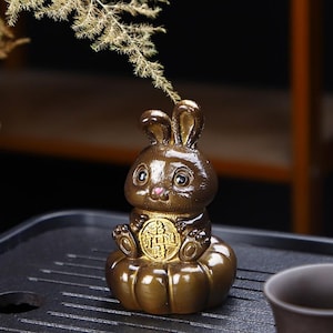 May include: A glossy dark brown ceramic rabbit figurine sitting on a pumpkin-shaped base. The rabbit has large eyes, a small pink nose, and holds a round gold-colored coin featuring Chinese characters in its front paws. A dried plant sprig is visible.