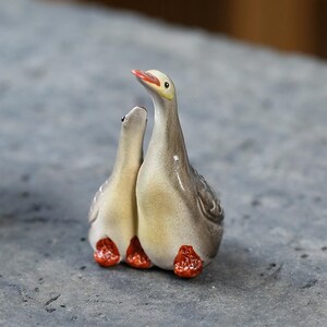Peut inclure: Ensemble de figurines en céramique représentant deux oies. Les oies ont des corps gris et crème avec des pieds orange et des becs rouges. L'une des oies regarde vers le haut, l'autre regarde la première oie.