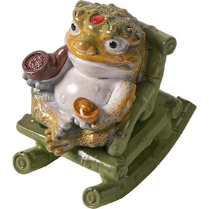 May include: A ceramic frog figurine with a red gem on its head, holding a pipe and a gold coin, seated on a green bamboo rocking chair. The frog is primarily yellow and brown with white accents.