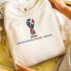 May include: Cream-colored sweatshirt featuring the 2026 FIFA World Cup logo. The logo is a stylized design with red, green, blue, and white elements. The text "2026 UNITED STATES CANADA MEXICO" is printed below the logo.