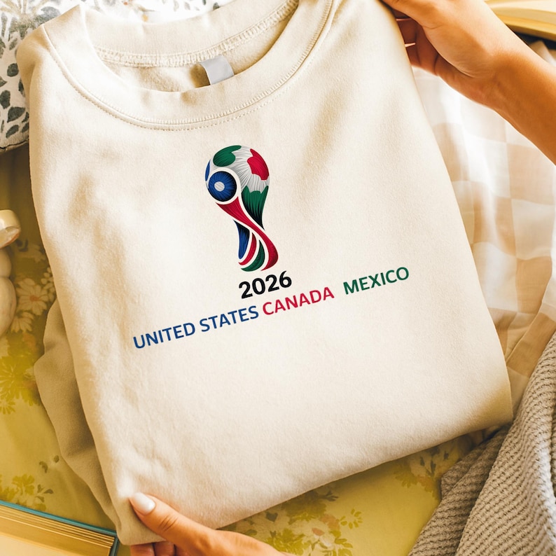 May include: Cream-colored sweatshirt featuring the 2026 FIFA World Cup logo. The logo is a stylized design with red, green, blue, and white elements. The text "2026 UNITED STATES CANADA MEXICO" is printed below the logo.