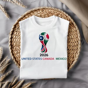 May include: White t-shirt featuring a colorful graphic of a soccer ball design with the text "2026" and "UNITED STATES CANADA MEXICO" in a bold font. The shirt is displayed on a woven basket.