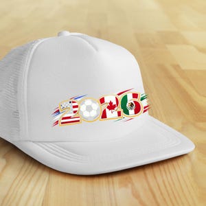 Puede incluir: Gorra de camionero blanca con visera curva. La parte delantera muestra el n&uacute;mero "2026" con un bal&oacute;n de f&uacute;tbol y las banderas de Estados Unidos, Canad&aacute; y M&eacute;xico. La gorra tiene una parte trasera de malla y una parte delantera s&oacute;lida.