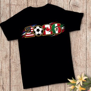 Puede incluir: Camiseta negra con la palabra "2026" en un dise&ntilde;o tem&aacute;tico de f&uacute;tbol. Los n&uacute;meros est&aacute;n estilizados con las banderas de Estados Unidos, Canad&aacute; y M&eacute;xico, y un bal&oacute;n de f&uacute;tbol en el centro.