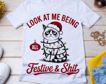 May include: White t-shirt featuring a grumpy cat wearing a Santa hat and Christmas lights. The text reads "Look at me being all festive & shit" in red script. A humorous holiday-themed graphic tee.
