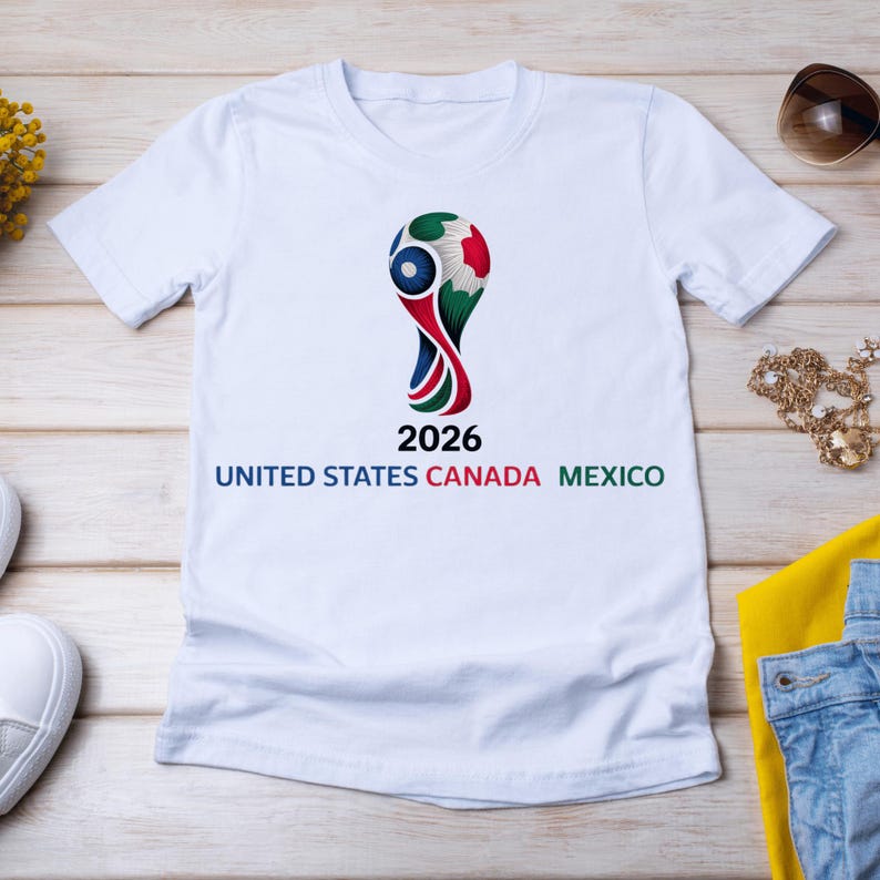May include: White t-shirt featuring a colorful graphic of a soccer ball trophy with the text "2026 UNITED STATES CANADA MEXICO". The design incorporates the colors of the flags of the three countries.