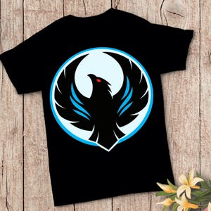 Peut inclure: T-shirt noir avec un motif de phénix stylisé. Le phénix est noir avec des accents bleus et un œil rouge, encadré dans un cercle bleu clair et blanc. Le motif est centré sur le t-shirt.