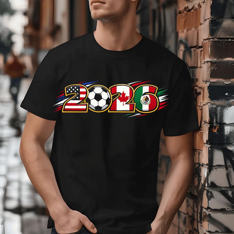 Puede incluir: Camiseta negra con el n&uacute;mero "2026" y un bal&oacute;n de f&uacute;tbol. El dise&ntilde;o incorpora las banderas de Estados Unidos, Canad&aacute; y M&eacute;xico, sugiriendo un tema deportivo.