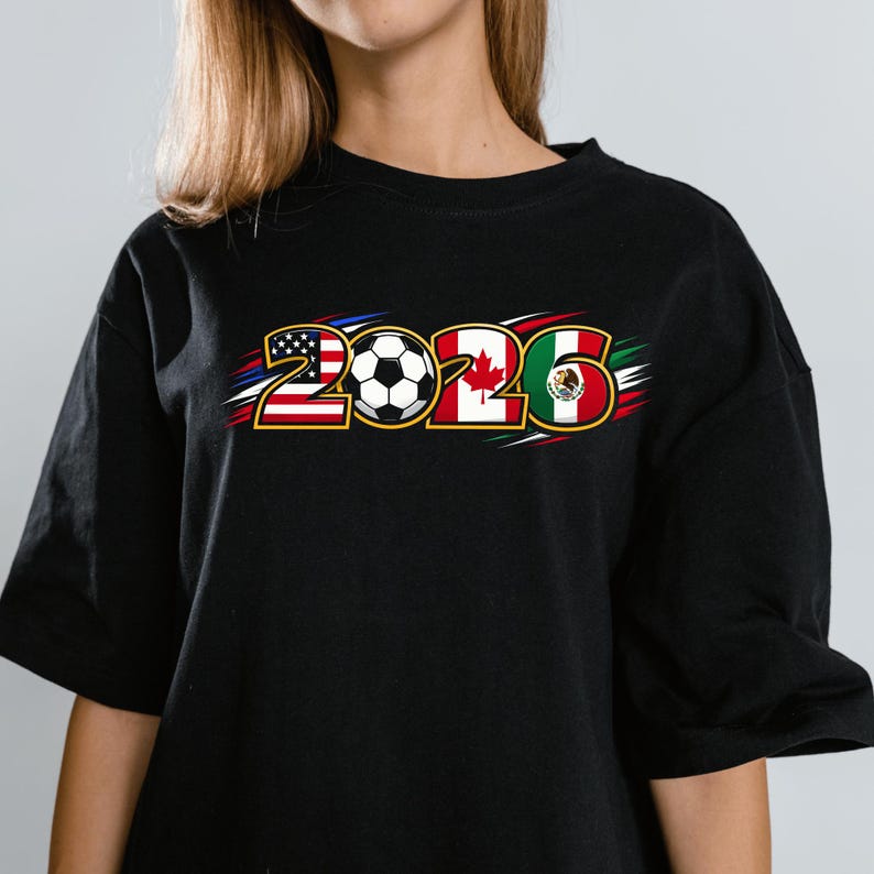 Puede incluir: Camiseta negra con el texto "2026" y un bal&oacute;n de f&uacute;tbol. Los n&uacute;meros est&aacute;n dise&ntilde;ados con las banderas de Estados Unidos, Canad&aacute; y M&eacute;xico, con detalles en rojo, blanco y azul.