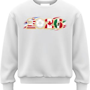 Puede incluir: Sudadera blanca con el n&uacute;mero "2026" que incorpora las banderas de Estados Unidos, Canad&aacute; y M&eacute;xico, junto con un gr&aacute;fico de bal&oacute;n de f&uacute;tbol. El dise&ntilde;o tiene detalles de pinceladas rojas, blancas y azules.