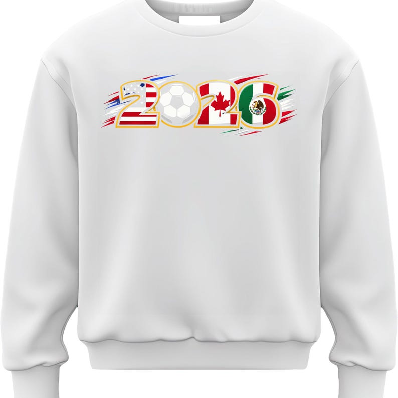 Puede incluir: Sudadera blanca con el n&uacute;mero "2026" que incorpora las banderas de Estados Unidos, Canad&aacute; y M&eacute;xico, junto con un gr&aacute;fico de bal&oacute;n de f&uacute;tbol. El dise&ntilde;o tiene detalles de pinceladas rojas, blancas y azules.