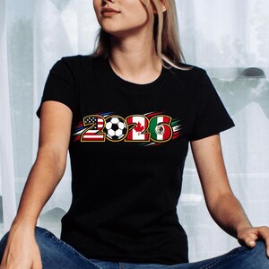Puede incluir: Camiseta negra con el a&ntilde;o "2026", un bal&oacute;n de f&uacute;tbol y las banderas de Estados Unidos, Canad&aacute; y M&eacute;xico. El dise&ntilde;o tiene detalles en rojo, blanco y azul.