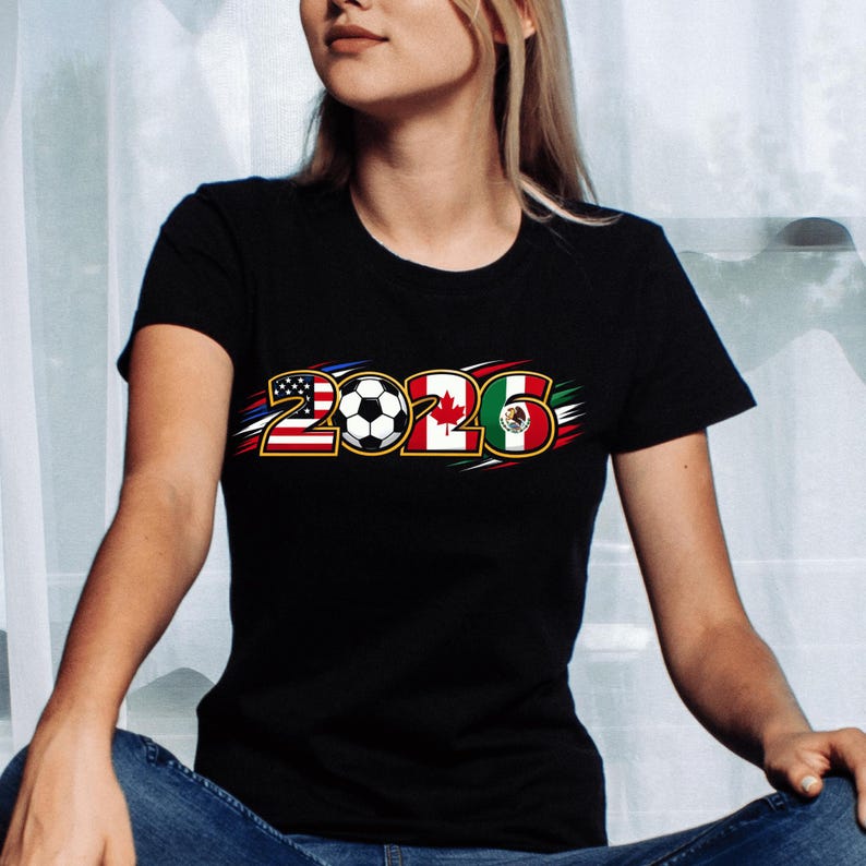 Puede incluir: Camiseta negra con el a&ntilde;o "2026", un bal&oacute;n de f&uacute;tbol y las banderas de Estados Unidos, Canad&aacute; y M&eacute;xico. El dise&ntilde;o tiene detalles en rojo, blanco y azul.