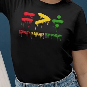 May include: Black t-shirt with a graphic design. The design features the words "EQUALITY IS GREATER THAN DIVISION" in red, yellow, and green dripping paint. The design also includes the symbols for equals, greater than, and division.