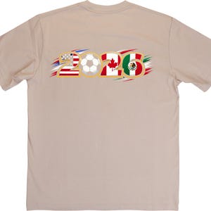 Puede incluir: Camiseta color beige con el texto "2026" en dorado. El n&uacute;mero "2" incorpora la bandera estadounidense, un bal&oacute;n de f&uacute;tbol en el centro, el segundo "2" tiene la bandera canadiense y el "6" la bandera mexicana. Los acentos son rojos, blancos, azules, verdes y dorados.