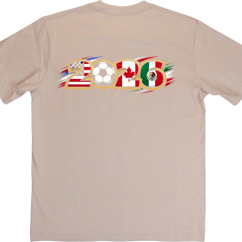 Puede incluir: Camiseta color beige con el texto "2026" en dorado. El n&uacute;mero "2" incorpora la bandera estadounidense, un bal&oacute;n de f&uacute;tbol en el centro, el segundo "2" tiene la bandera canadiense y el "6" la bandera mexicana. Los acentos son rojos, blancos, azules, verdes y dorados.