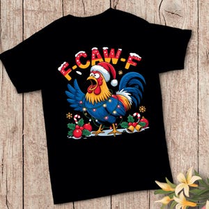 Puede incluir: Camiseta negra con un gallo de dibujos animados que lleva un gorro de Papá Noel y luces navideñas. El gallo tiene plumas azules y una cresta roja. El texto "F-CAW-F" está encima del gallo en una fuente festiva. Acebos y bastones de caramelo completan el diseño.