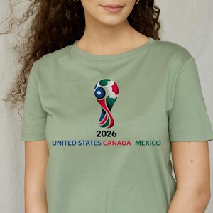 May include: Sage green t-shirt featuring a colorful graphic of a soccer trophy with the text "2026" and "UNITED STATES CANADA MEXICO" in a bold font. The shirt is a solid color.
