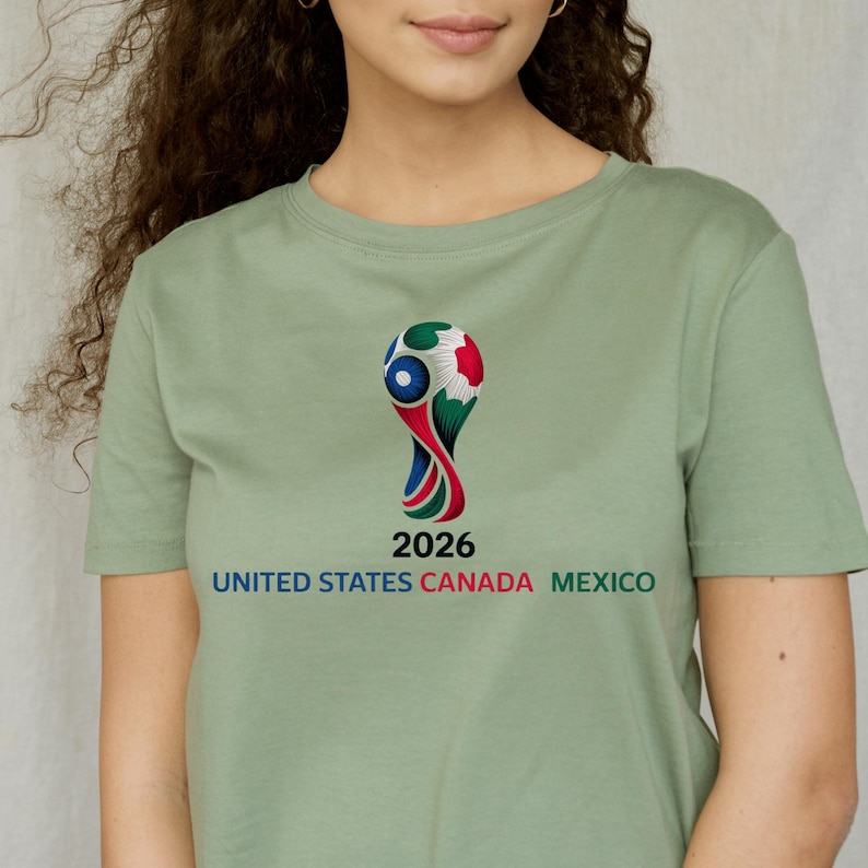 May include: Sage green t-shirt featuring a colorful graphic of a soccer trophy with the text "2026" and "UNITED STATES CANADA MEXICO" in a bold font. The shirt is a solid color.