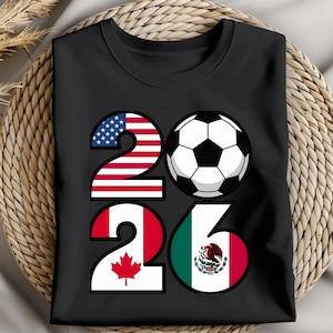 May include: Black t-shirt featuring the number "26" with the flags of the United States, Canada, and Mexico. A soccer ball graphic is between the numbers. The shirt is folded on a woven surface.