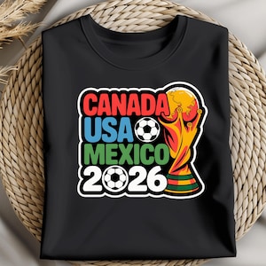 以下が含まれることがあります： 2026 FIFAワールドカップのカラフルなグラフィックがプリントされた黒いTシャツ。「CANADA USA MEXICO 2026」の文字が赤、青、緑でデザインされ、サッカーボールとトロフィーが描かれています。