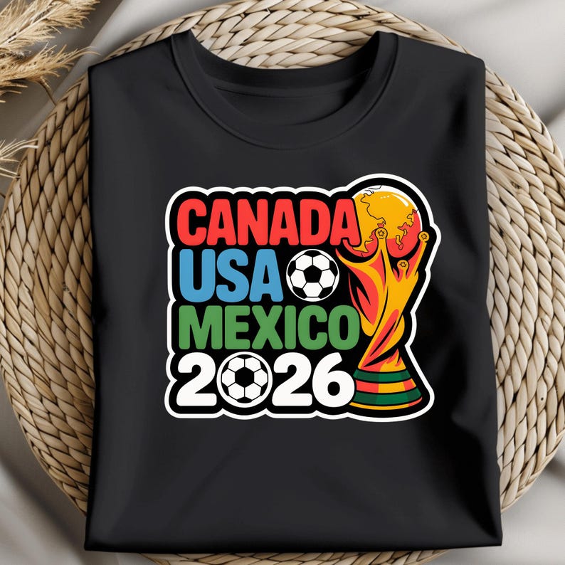 以下が含まれることがあります： 2026 FIFAワールドカップのカラフルなグラフィックがプリントされた黒いTシャツ。「CANADA USA MEXICO 2026」の文字が赤、青、緑でデザインされ、サッカーボールとトロフィーが描かれています。